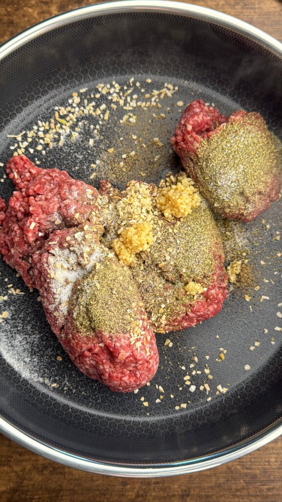 Ground beef in a frying pan topped with minced garlic, dried onions, black pepper, and salt, ready to be cooked.