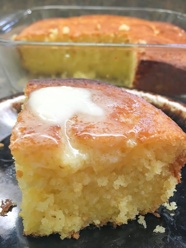 A close-up of a moist slice of cornbread with melted butter on top, placed on a plate, with the remaining cornbread loaf in the background.