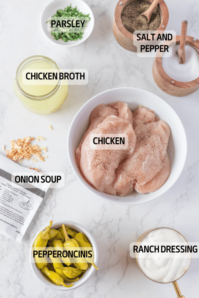 A marble counter displays labeled ingredients for a recipe: raw chicken breasts in a bowl, chicken broth, onion soup mix, pepperoncinis, ranch dressing, chopped parsley, and bowls of salt and pepper.
