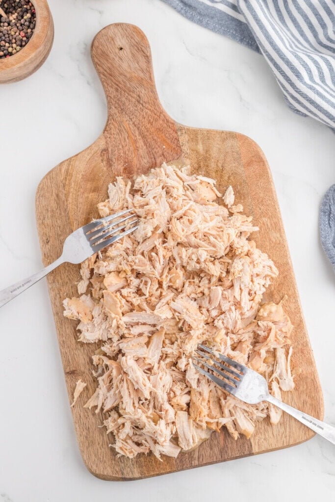 Shredded cooked chicken sits on a wooden cutting board with two forks used for pulling apart the meat. A bowl of pepper and a striped cloth are nearby on a white surface.