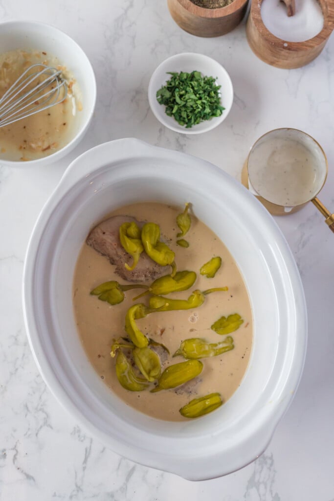 A white slow cooker filled with meat, creamy sauce, and sliced pepperoncini peppers sits on a marble surface. Surrounding it are bowls of chopped herbs, seasoning, and a whisk in a small bowl of sauce.