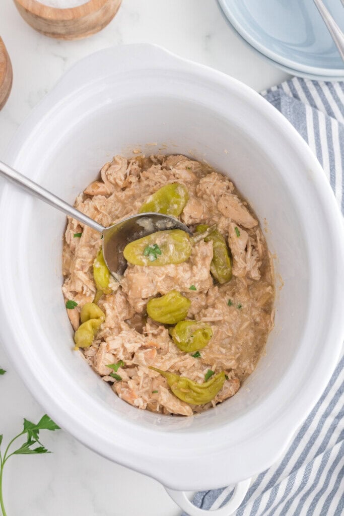 A white slow cooker filled with shredded chicken, creamy sauce, and whole pepperoncini peppers, with a serving spoon inside. The slow cooker rests on a white countertop next to a striped kitchen towel.