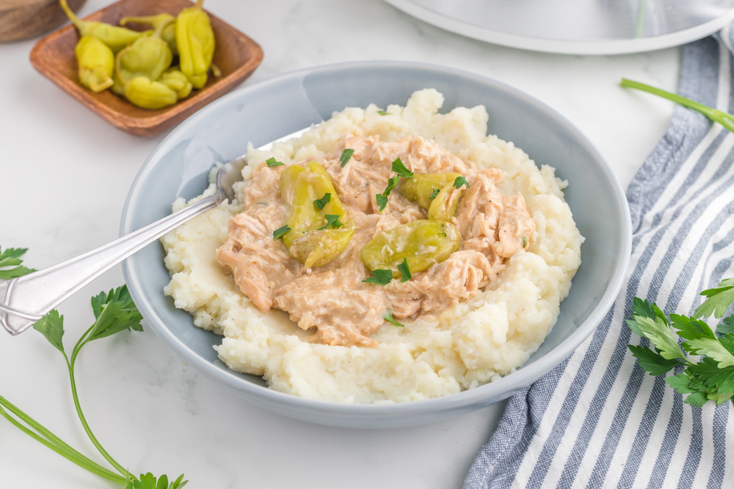 A bowl of mashed potatoes topped with creamy shredded chicken and sliced pepperoncini peppers, garnished with parsley. A fork rests on the side, with fresh parsley and a striped napkin nearby.