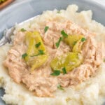 A plate of mashed potatoes topped with shredded chicken in a creamy sauce, garnished with whole pepperoncini peppers and chopped parsley.
