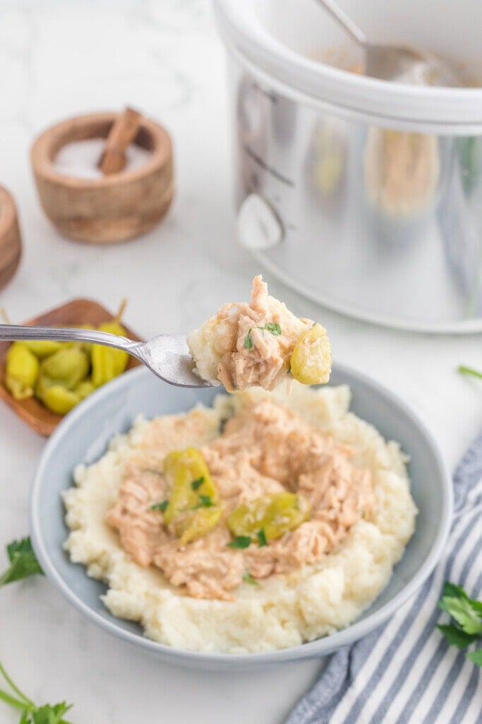 A bowl of mashed potatoes topped with shredded creamy chicken and sliced peppers. A fork holding a bite of the dish is in focus, with a slow cooker and small bowls of ingredients in the background.