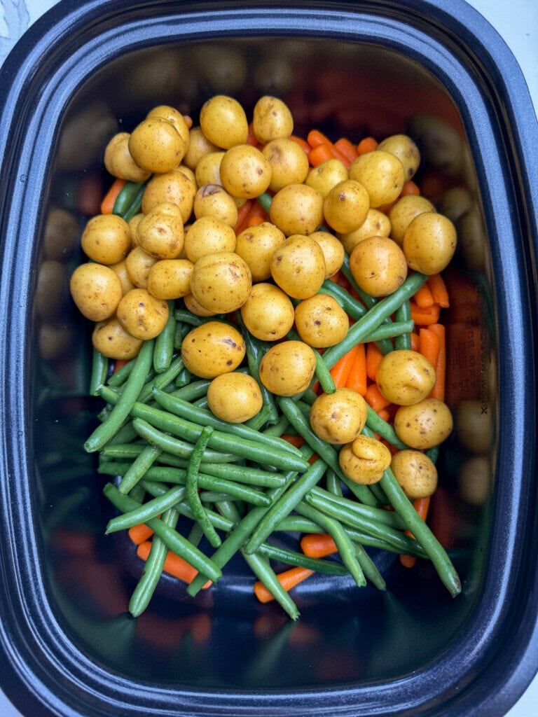 A slow cooker filled with small yellow potatoes, fresh green beans, and baby carrots, all uncooked and layered inside the pot.