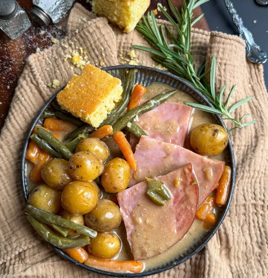 A bowl of ham slices with baby potatoes, carrots, and green beans in gravy, garnished with rosemary, sits on a tan cloth. A square of cornbread rests near the bowl.