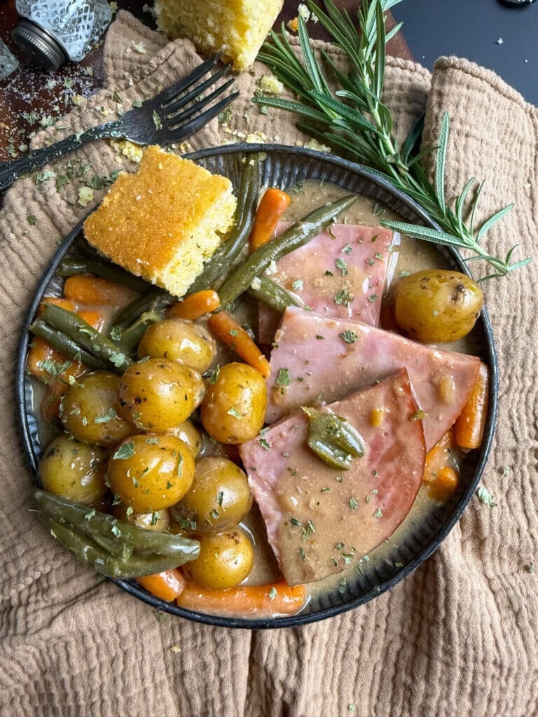 A plate with ham slices, baby potatoes, green beans, and carrots in gravy, garnished with herbs. A piece of cornbread and a sprig of rosemary are on the side, with a fork and brown napkin nearby.