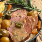 A plate with slices of glazed ham topped with gravy, surrounded by baby potatoes, carrots, green beans, and garnished with fresh rosemary. A piece of cornbread is visible in the background.