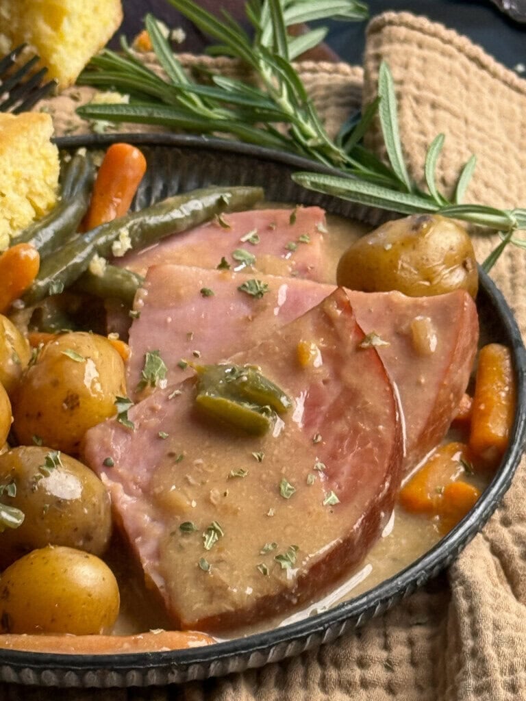 A plate with slices of glazed ham topped with gravy, surrounded by baby potatoes, carrots, green beans, and garnished with fresh rosemary. A piece of cornbread is visible in the background.