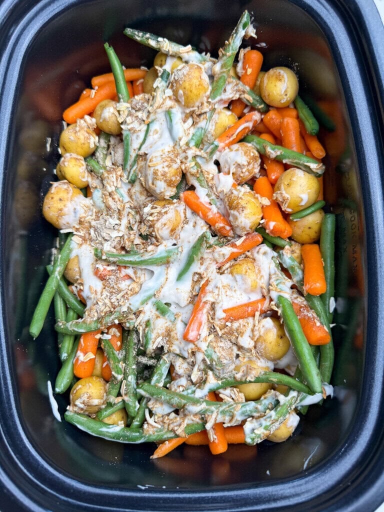 A slow cooker filled with baby potatoes, green beans, and baby carrots, topped with a creamy white sauce and crispy fried onions.