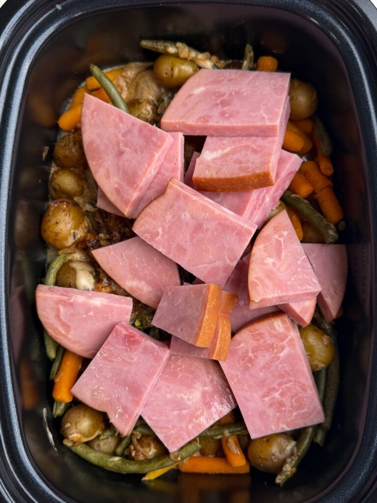 Sliced ham placed on top of a mix of baby potatoes, green beans, and carrot pieces inside a slow cooker or roasting pan.
