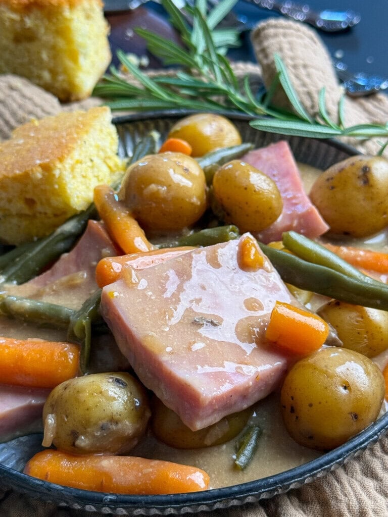 A close-up of a plate with slices of ham, baby potatoes, green beans, and carrots in a savory sauce, garnished with a sprig of rosemary. Cornbread pieces are visible in the background.