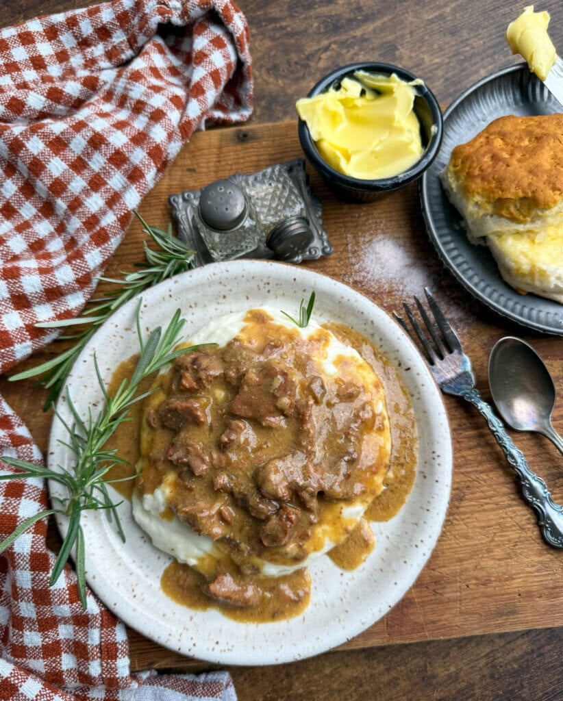 A plate of creamy mashed potatoes topped with beef gravy sits on a wooden table, garnished with rosemary. Nearby are a biscuit on a small plate, a bowl of butter, cutlery, and a red-checked cloth napkin.
