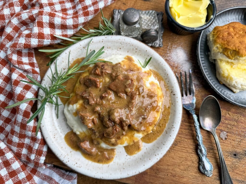 A plate of creamy mashed potatoes topped with beef gravy and a sprig of rosemary, beside a biscuit with butter on a small dish, a fork and spoon, salt and pepper shakers, and a red plaid napkin on a wooden table.