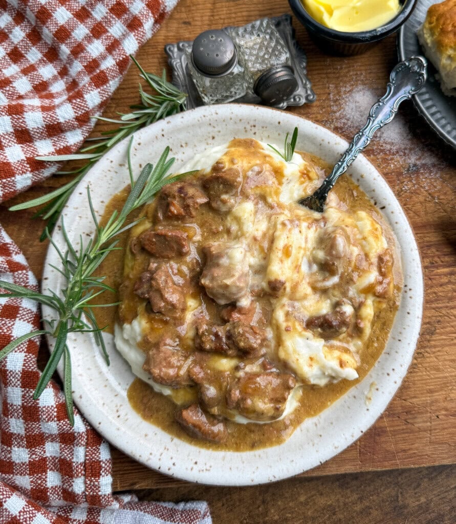 A plate of creamy mashed potatoes topped with beef gravy and chunks of beef, garnished with fresh rosemary. A fork rests on the plate. A checkered napkin, a butter dish, and salt and pepper shakers are nearby.
