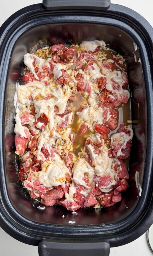 Raw beef chunks, cream of mushroom soup, dry onion soup mix, and broth are layered in a black slow cooker, ready to be cooked.