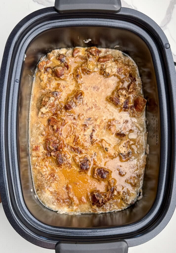 A slow cooker filled with cooked, saucy pieces of meat in a brown, oily broth. The dish appears tender and juicy, with small chunks and a glossy surface. The slow cooker sits on a white countertop.