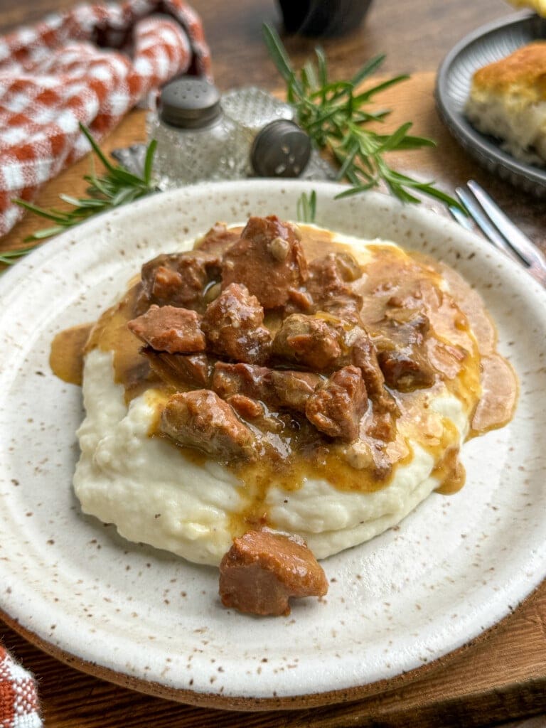 A plate of creamy mashed potatoes topped with savory beef tips in brown gravy, garnished with fresh rosemary, sits on a speckled white plate with a fork nearby.