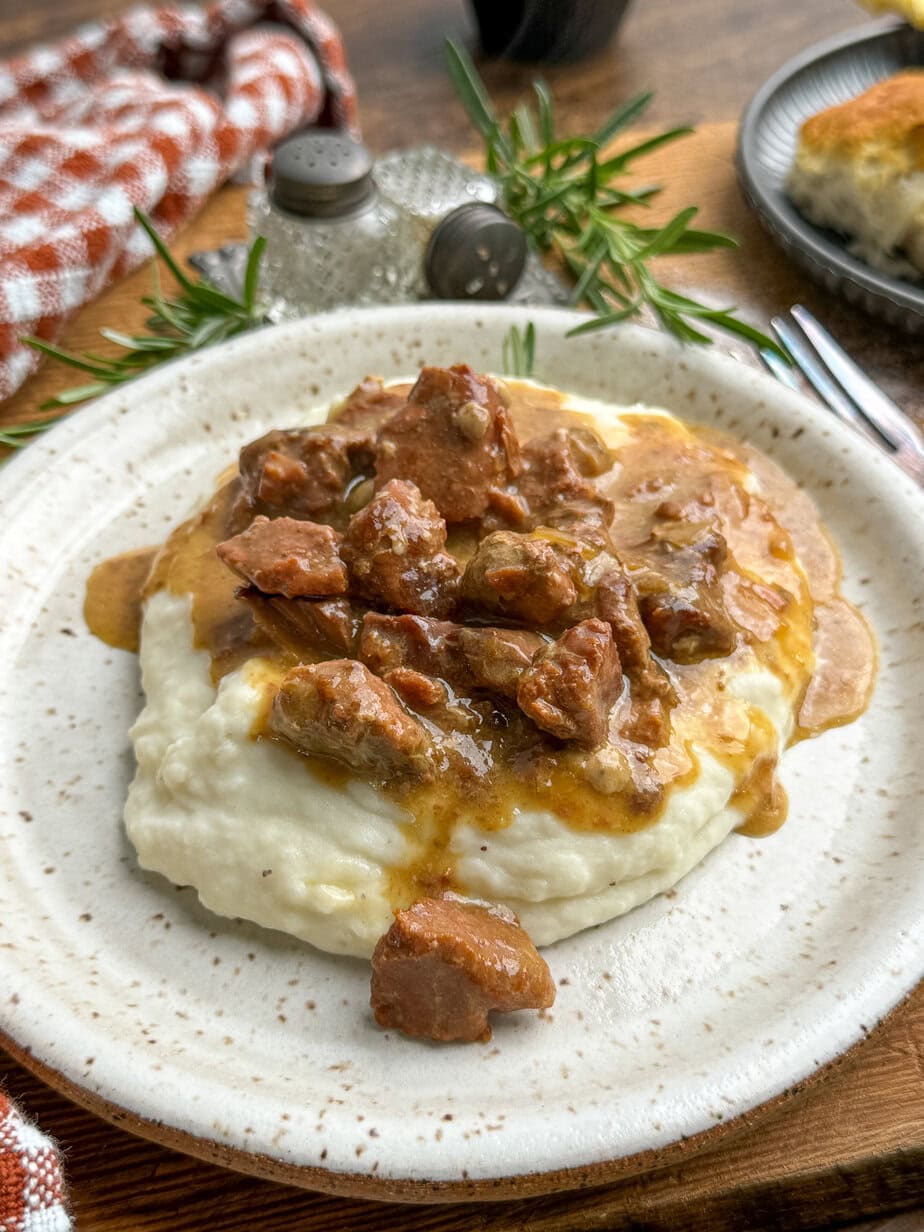 A plate of creamy mashed potatoes topped with savory beef tips in brown gravy, garnished with fresh rosemary, sits on a speckled white plate with a fork nearby.