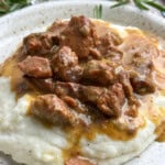 A plate of creamy mashed potatoes topped with tender beef stew in gravy, garnished with fresh rosemary. Salt and pepper shakers are visible in the background.
