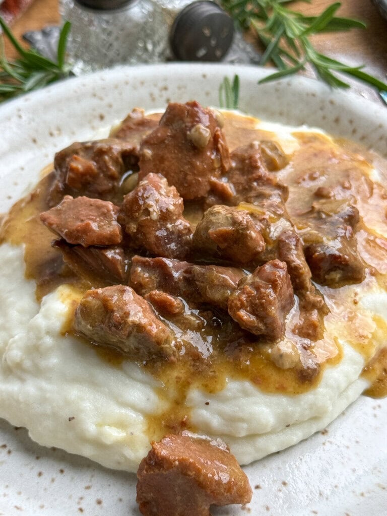 A plate of creamy mashed potatoes topped with tender beef stew in gravy, garnished with fresh rosemary. Salt and pepper shakers are visible in the background.