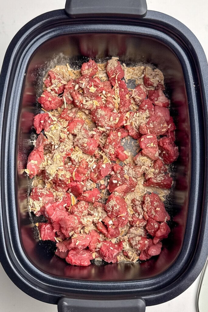 Chunks of raw beef and dried seasonings are spread in the bottom of a black slow cooker insert, ready to be cooked.