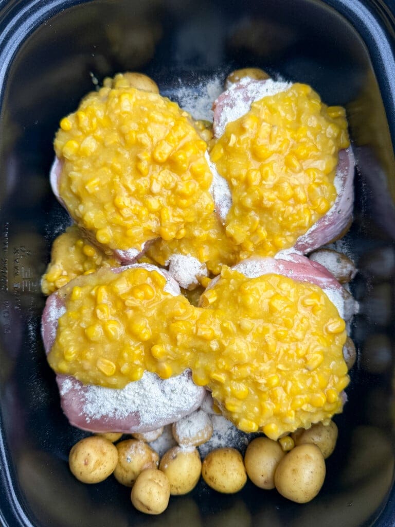 Raw pork chops in a slow cooker, topped with a creamy yellow corn mixture, with small whole potatoes arranged around the meat.