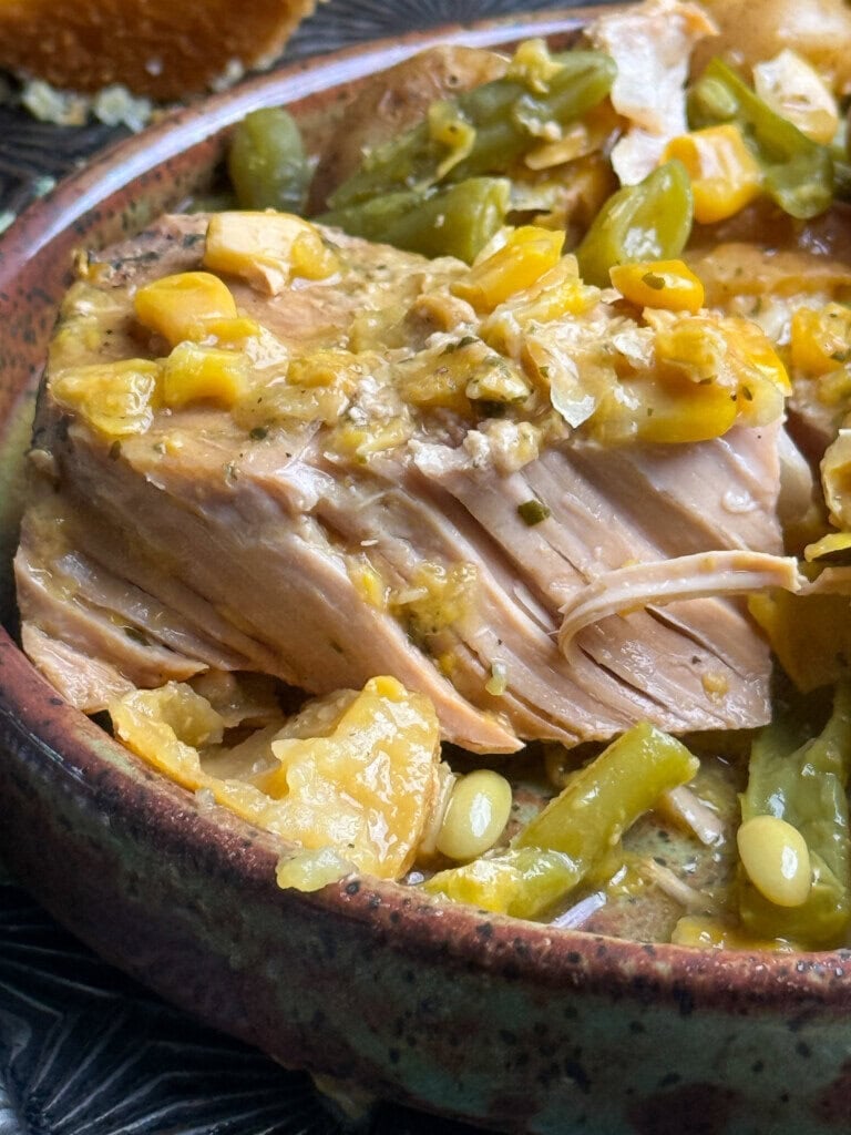 Close-up of a plate filled with tender, shredded pork, yellow corn, green beans, and a thick, seasoned broth. The food appears hearty and homemade, served in a rustic ceramic dish.