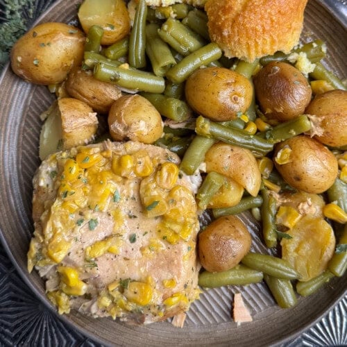 A plate of food with pork chops topped with corn, baby potatoes, green beans, and a piece of cornbread muffin.