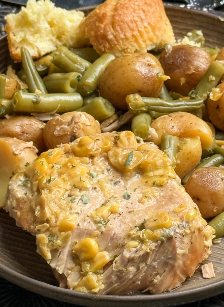 A plate of food featuring a piece of seasoned pork, green beans, small potatoes, and a piece of cornbread. The meal appears hearty and home-cooked, with a light gravy or sauce over the chicken.