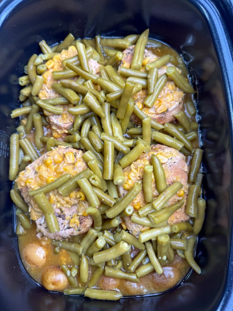 Pork chops topped with cooked green beans and corn in a brown sauce, served in a black rectangular slow cooker or casserole dish.