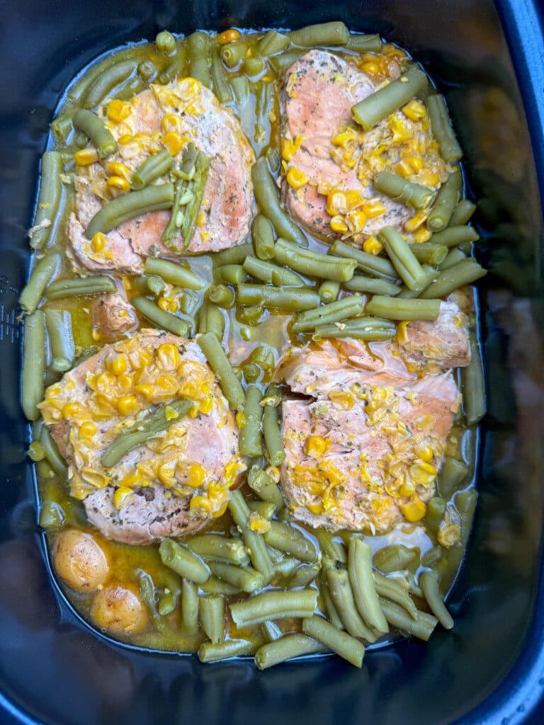 Four cooked pork chops topped with corn and surrounded by green beans and a few small potatoes in a dark rectangular dish, covered in a yellowish broth or sauce.