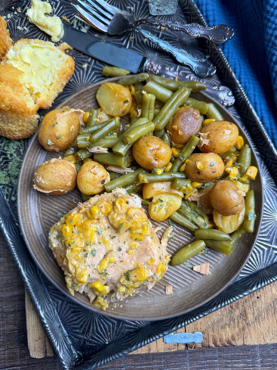 A plate with seasoned pork chops topped with corn, green beans, baby potatoes, and sliced carrots, served with buttered cornbread. Vintage cutlery and a blue napkin are next to the plate on a decorative tray.