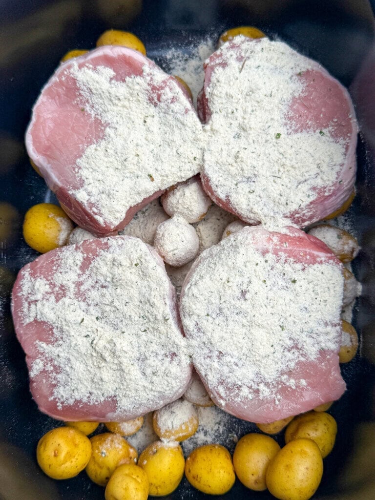 Four raw pork chops coated with a layer of ranch seasoning rest on top of whole baby potatoes in a black cooking pot, ready to be cooked.