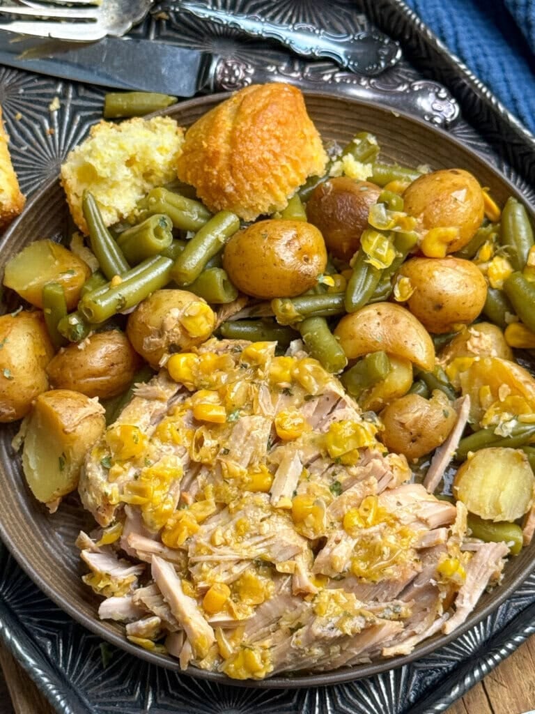 A plate filled with pulled pork topped with corn, roasted baby potatoes, green beans, and pieces of cornbread, served on a decorative metal tray with a fork and knife.