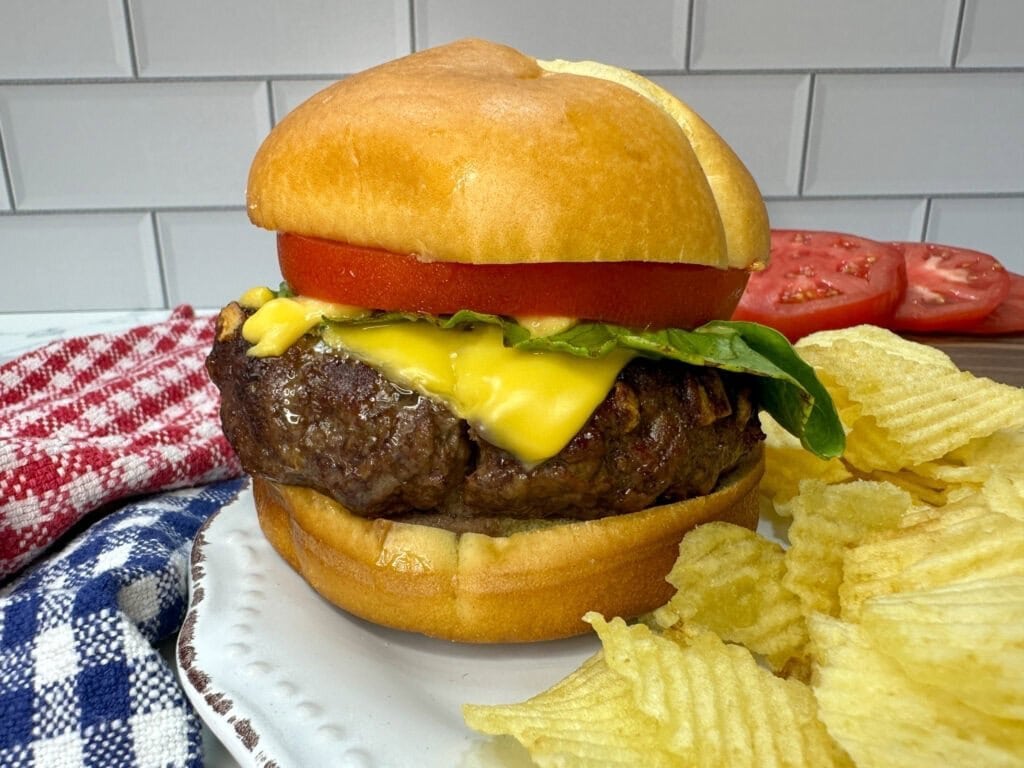 A cheeseburger with lettuce, tomato, and melted cheese on a bun sits on a white plate next to a pile of potato chips. A red and white checked napkin and sliced tomatoes are in the background.
