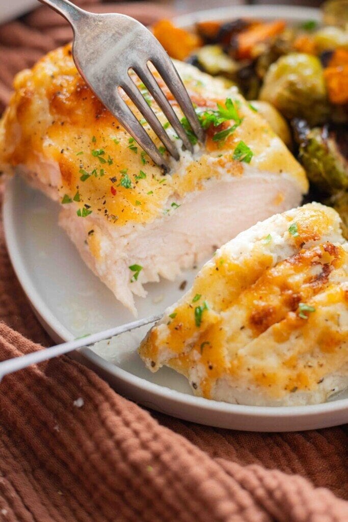 A close-up of a fork cutting into a juicy, baked chicken breast topped with melted cheese and herbs, served with roasted vegetables on a white plate—perfect for anyone following How to Cook for One Person: The Complete Guide to Real Dinners Without the Waste.