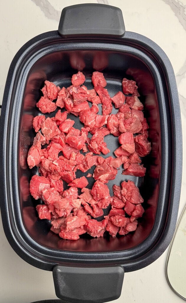 Diced raw beef pieces spread evenly in the bottom of a black slow cooker on a light-colored countertop.