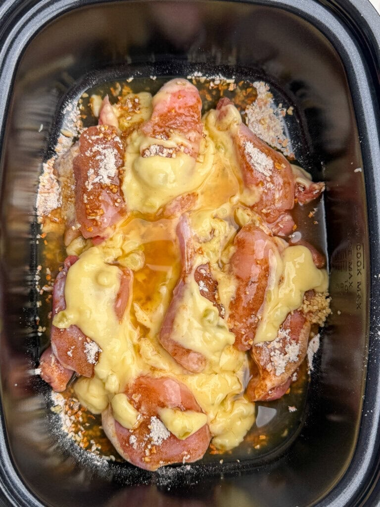 Chicken thighs in a slow cooker, topped with a creamy yellow sauce, seasonings, and what appears to be breadcrumbs or grated cheese, ready to be cooked.