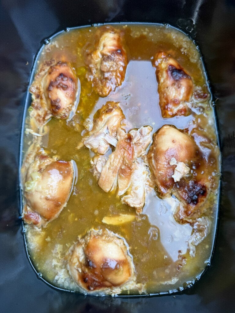 Several pieces of cooked chicken in a brown, saucy liquid inside a black slow cooker or crockpot. The chicken appears tender and partially shredded.