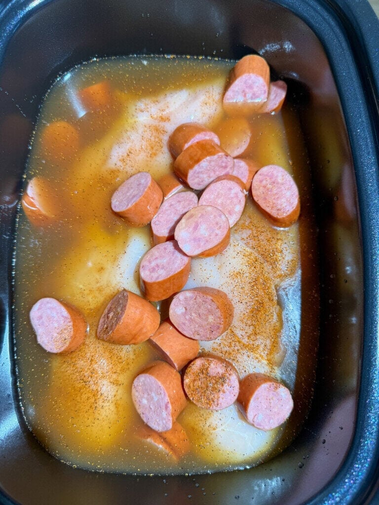Sliced sausages and seasoned liquid in a slow cooker, preparing to cook along with chicken breasts submerged in the broth.