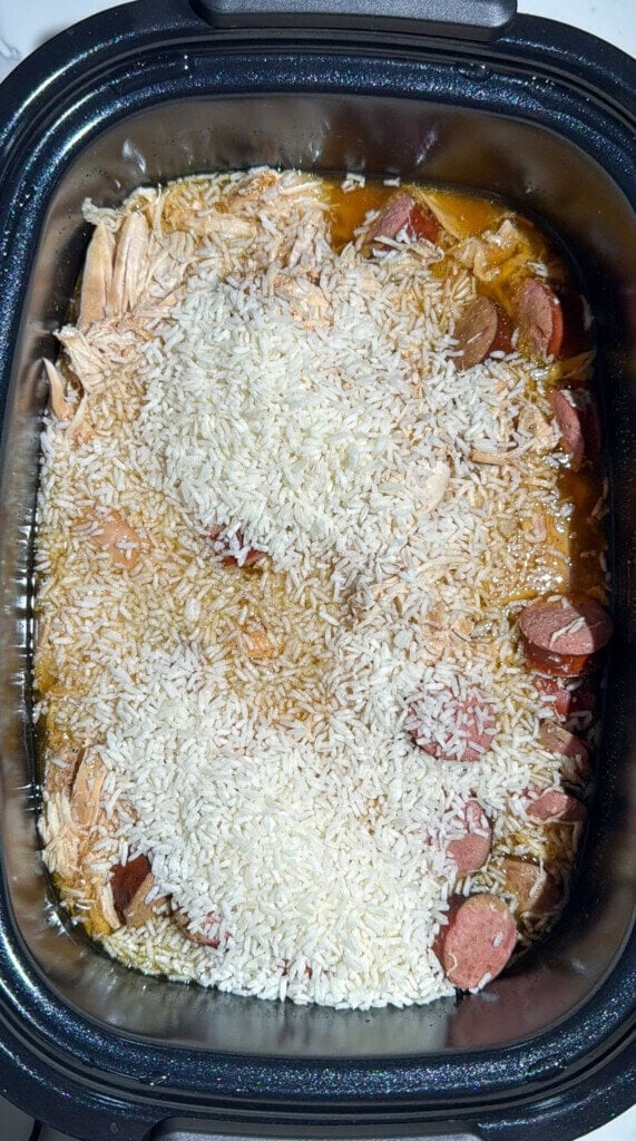 A close-up of a slow cooker filled with shredded chicken, sliced sausage, uncooked white rice, and broth, ready to be cooked.
