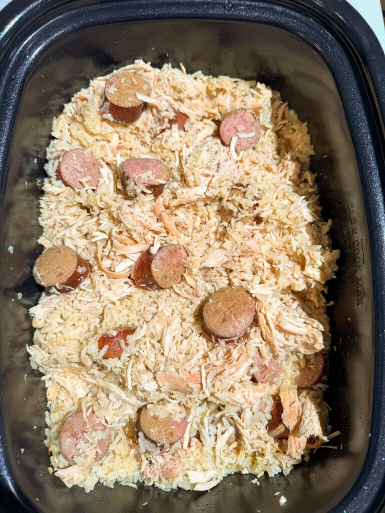 Shredded chicken, sliced sausage, and rice mixed together in a black slow cooker, ready to be cooked or served.