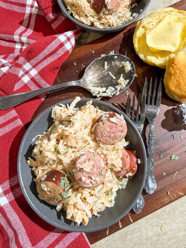 A plate of creamy shredded chicken and sausage garnished with herbs sits next to a fork, spoon, and a buttered roll on a wooden board with a red plaid napkin.