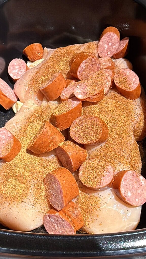 Raw chicken breasts and sliced sausage are placed in a black pan, sprinkled generously with a reddish-orange seasoning blend.