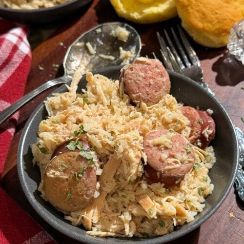A bowl of shredded chicken and sliced sausage mixed with rice and herbs, served on a wooden table next to a buttered biscuit and a red-and-white checkered napkin. A spoon and fork are nearby.