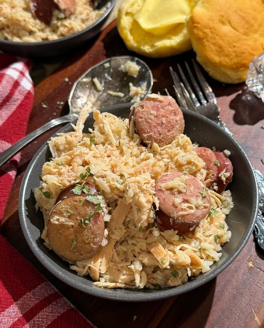 A bowl of shredded chicken and sliced sausage mixed with rice and herbs, served on a wooden table next to a buttered biscuit and a red-and-white checkered napkin. A spoon and fork are nearby.