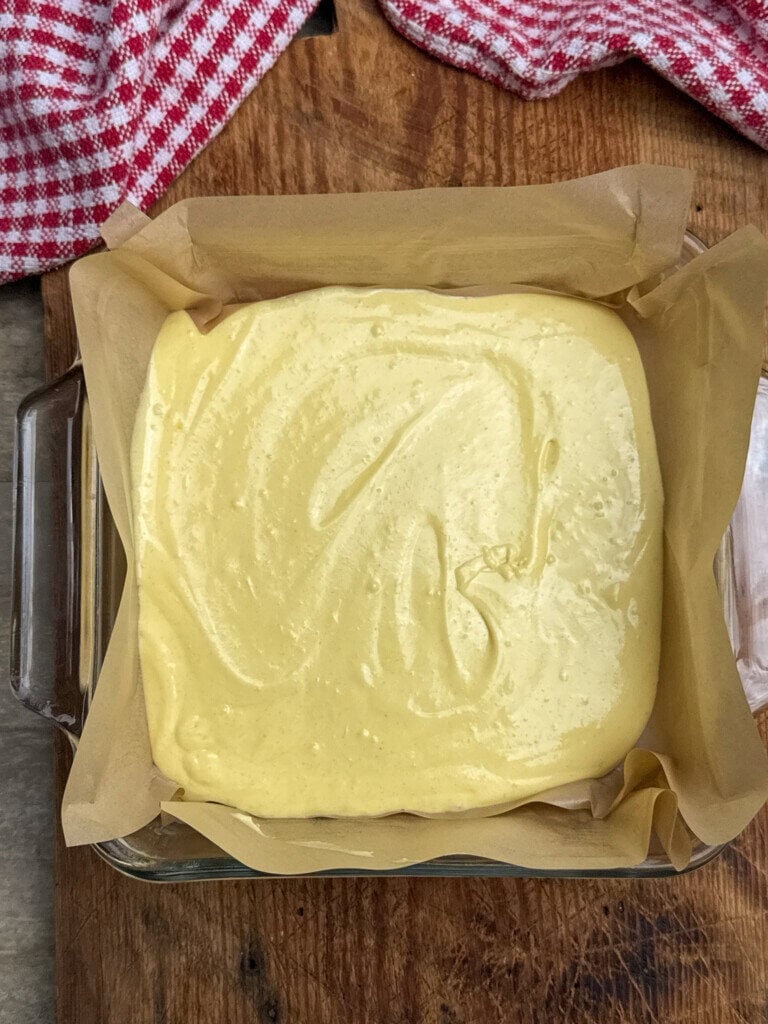 A square glass baking dish lined with parchment paper filled with smooth, uncooked yellow cake batter sits on a wooden surface, with a red and white checkered cloth nearby.