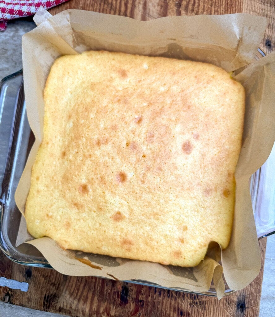 A freshly baked square cake with a golden-brown top sits on brown parchment paper in a glass baking dish on a wooden surface.
